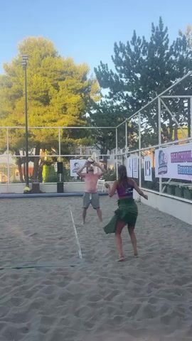 A man and a woman are playing beach volleyball on a sand court. The man is shirtless and wearing grey shorts, and he is in the process of hitting a volleyball. The woman is wearing a purple sports bra and a green skirt, and she is facing away from the camera, with her arms raised as if she is about to receive the ball. There is a net between them, and a fence behind the court. There are trees and a building in the background. There are also several banners and signs visible behind the fence. The lighting suggests it is daytime, possibly late afternoon due to the warm lighting.