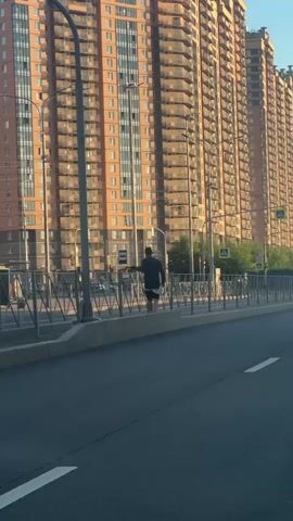 A person is skateboarding near a tall apartment building. The person is wearing a dark shirt and shorts. The skateboard is black. The person is on a sidewalk next to a road. There are trees and streetlights in the background. The sun is shining.
