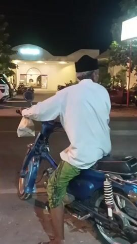A man wearing a white long-sleeved shirt, green patterned pants, and a black cap rides a blue motorcycle. He is facing away from the camera and holding onto the handlebars. The motorcycle has a silver rack on the back. In the background, there is a building with lights and signs, and trees. Another motorcycle passes by, carrying two people. The scene appears to be at night.