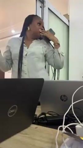 A woman with long braids wearing a light green blouse is standing indoors in what appears to be an office space. She has a surprised expression and her hand is near her mouth, as if gasping. She is holding a phone to her ear. In front of her on a desk are two laptops, one a Dell and the other an HP. Various cables and power adapters are also visible on the desk. A glass door or window is behind her. The room is well-lit.