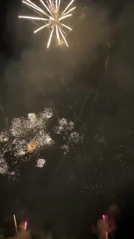 The video captures a fireworks display at night. Multiple fireworks explode in the dark sky, emitting bright bursts of light and trailing sparks. Some fireworks create a starburst effect, while others form a cascade of sparks. Smoke trails from the explosions, partially obscuring the view of the sky. The overall scene is illuminated by the light of the fireworks, creating a festive and dynamic atmosphere. There are no people or specific objects visible in the frame, only the fireworks and the night sky.