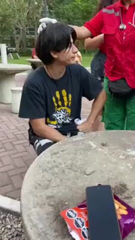 A young man with long dark hair and glasses, wearing a black t-shirt, patterned shorts, and black shoes, sits at a round stone table in an outdoor park setting. His left hand is clenched. An older man in a red shirt sits at another table in the background. A woman in a red top and another person in a red jacket with green pants stand near the young man, seemingly attending to a possible head injury. The person in the red jacket is wearing white gloves. On the table near the young man is a smartphone and opened snacks. The park features brick pathways, stone tables, benches, and trees.