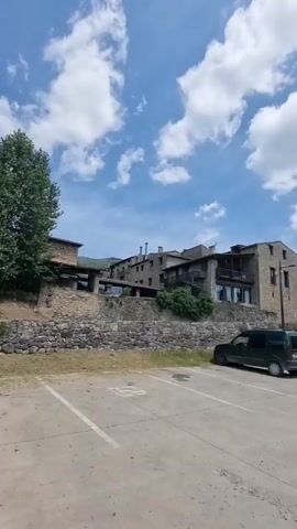 This media file captures a daytime scene in Montagut i Oix, Spain. The foreground features a paved parking lot with several vehicles, including a dark green van, a light-colored SUV, and what appears to be a utility trailer. Beyond the parking lot, a stone wall separates it from a hillside covered in buildings. These structures are made of stone and have a rustic, traditional appearance, with visible chimneys and balconies. One building on the right side of the cluster has a bell tower, suggesting it might be a church or a similar public building. The sky above is a vibrant blue, dotted with fluffy white clouds, indicating fair weather. The overall setting is a village nestled in a hilly or mountainous region, as suggested by the distant green slopes visible in the background. The time of day appears to be midday or early afternoon due to the bright sunlight casting shadows. There is no visible text within the media file.