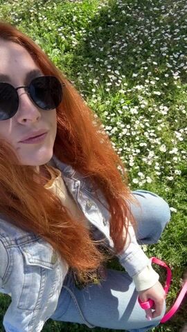 A woman with long red hair, wearing sunglasses and a denim jacket, sits on a grassy field dotted with small white flowers, holding a pink leash attached to a small, grey dog. The dog walks around a black, vertical object. The background includes a wire fence and a sunny sky.