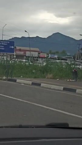 The video shows a roadside view with a large mountain in the background under a cloudy sky. In the foreground, there is a paved road with a black and white striped curb. Adjacent to the curb is a patch of tall green grass, and beyond that, several parked white vehicles. A few commercial buildings are visible behind the vehicles, some with signage. To the left, a blue directional sign with white text and an arrow is mounted on a pole. Further to the right, a black trash can stands near the edge of the grassy area, with some litter scattered around it. A dark-colored sedan is partially visible moving along the road, with its roof rack clearly distinct. The overall impression is a somewhat overcast day in a semi-urban or roadside setting near a mountainous region.