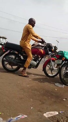 A man in a tan outfit sits on a red and black motorcycle parked on a weathered, dirty street. He is wearing sandals. Other motorcycles are parked nearby. There is litter scattered on the ground. Another man walks by the parked motorcycles. The sky is overcast and power lines are visible overhead.