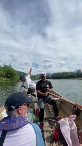 A group of four people are on a long, narrow wooden boat being rowed down a river. The boat is moving forward. The sky is cloudy and the sun is visible. The riverbanks are lined with trees and vegetation. Two men are in the back of the boat, rowing. One man is in the middle of the boat, holding a paddle. One person is in the front of the boat, facing forward. The camera is positioned in the front of the boat, looking back at the people. The video is shot from a first-person perspective.