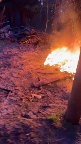 The video shows a person in dark clothing tending a large bonfire. The person uses a stick to stir the flames. Sparks fly upwards from the fire. The camera pans to show a wooden gazebo-like structure nearby, with other people sitting inside. The environment appears to be a wooded area with a sandy ground. The light suggests it is dusk or evening.