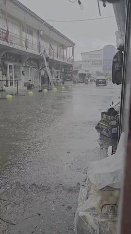 The video shows a street scene during a heavy rainstorm. Water is pooling on the ground, creating reflections of the buildings and objects around it. In the background, a person is on a ladder, possibly working on one of the buildings. A few cars are visible driving down the flooded street. Yellow spherical objects are scattered along the sidewalk. In the foreground, there are some items covered in plastic sheeting, suggesting they are being protected from the rain. The sky is overcast and grey, consistent with a downpour. The overall impression is one of disruption and inconvenience caused by the weather.