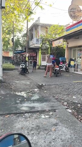 The video shows a street scene with several people and vehicles. In the foreground, there's a somewhat dilapidated street with puddles and debris. A motorcycle is visible in the bottom left corner, presumably where the camera is positioned.

The middle ground features a paved area where two people are standing near a motorcycle with a rider on it. One person is wearing a headscarf and light-colored pants, while the other is wearing dark clothing. Another motorcycle with a rider is further to the left.

In the background, there's a building with an Indomaret sign, indicating a convenience store. Another person is seated on the ground in front of the store. Other buildings and trees are also visible, along with overhead wires. The lighting suggests it might be daytime, possibly late afternoon.