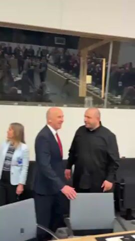 Here is a brief description of the video:

The video shows two men posing for the camera in what appears to be a break area or waiting room. The man on the left is wearing a dark suit with a red tie. The man on the right is wearing a dark button-down shirt. They are standing near a conference room visible through a glass wall. A group of people is present in the conference room. The break area is furnished with office chairs and tables.