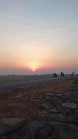 The video shows a sunset over a rural landscape. In the foreground, dry grass and rocks line a path. A paved road stretches into the distance, with a green field on the left and a hazy horizon on the right. A few motorcycles are visible on the road, with people riding them. The sun is a bright, pinkish-orange orb low in the sky, casting a warm glow over the scene. The sky is a gradient of pale yellow to pink and blue, with thin clouds. The overall mood is peaceful and serene.