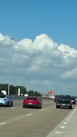 A vertical video captures a view from a vehicle traveling on a multi-lane highway. The sky is bright blue with large, puffy white clouds. Tall streetlights are visible along the side of the road. Several cars are on the highway, including a blue sports car, a red sports car, a black pickup truck, and a gray sedan. Some of the cars are in the foreground, while others are further down the road. There are orange construction barrels and signs visible on the right side of the highway, indicating a construction zone. The road appears to be in good condition, with clear lane markings. The overall scene suggests a sunny day with moderate traffic on a highway.