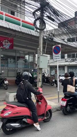 The video begins with a view of a street scene in what appears to be a city. In the foreground, a person wearing a black helmet and black clothing is seated on a pink scooter, facing left. To the right of the pink scooter, a black scooter carries two people, a male driver and a female passenger wearing a white shirt and carrying a light brown bag. Behind the black scooter, a gray car is partially visible.

The background shows a busy street with buildings, overhead electrical wires, and several parked motorcycles. A 7-Eleven store is visible on the left side of the frame. A blue and white street sign with Thai writing and a red circle with a diagonal line inside, indicating a no parking zone, is mounted on a pole. A man in a gray shirt is standing near the sign.

As the video progresses, the camera pans to the right, revealing more of the street traffic. The gray car moves forward, followed by the black scooter. More buildings and electrical wires dominate the upper portion of the frame. Further down the street, other vehicles, including a red truck and more cars, are visible, suggesting moderate traffic. The sky is overcast with clouds. A person wearing a bright green jacket and a black helmet is seen riding a motorcycle in the foreground, facing away from the camera. The video captures a snapshot of urban life, likely in Southeast Asia, with a focus on street-level activity and the presence of motorbikes as a common mode of transportation.