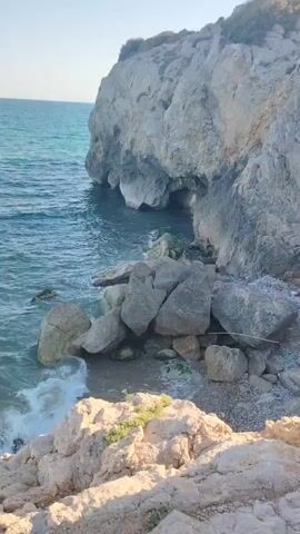 The video shows a natural coastal scene with rough waves crashing against large rock formations. The water is a deep blue and appears to be clear. The rocks are a mix of light and dark gray, some with patches of green algae. The scene is brightly lit by sunlight, suggesting it is daytime. The camera is positioned at a slight angle, looking down towards the water and rocks. There are no people or man-made objects visible in the frame.