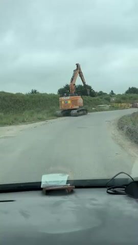 The video shows the view from inside a vehicle, looking out at a large orange excavator. The excavator is positioned on the side of a paved road, with its tracks on the grassy verge. The excavator's boom is raised and the bucket is extended, appearing to be in the process of digging or moving material. The sky is overcast and the environment appears to be a rural or construction site setting with trees and bushes in the background. The interior of the vehicle is partially visible in the foreground, including the dashboard and part of the windshield. There are some items on the dashboard, including a small stand with what appears to be a piece of paper or a small object, and some packets of snacks. The camera is moving forward, giving a sense of the vehicle's movement along the road.