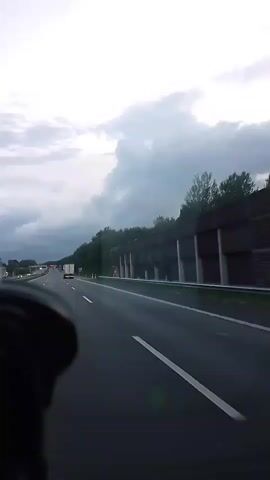 The video shows a view from the perspective of a driver on a highway. The road is wet, suggesting it may have rained recently or is raining lightly. Dark, heavy clouds dominate the sky. On the right side of the highway, there is a sound barrier or retaining wall covered in vegetation, and further on, a dense forest. On the left side of the highway, other vehicles, including a white truck, can be seen in the distance. The driver's steering wheel is partially visible in the lower left corner of the frame.
