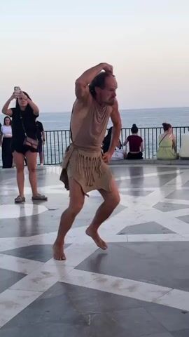 A barefoot man dressed in a tunic and loincloth performs a dramatic dance on a patterned stone floor. He has a stern expression and his arms are raised. In the background, a railing separates the performance area from a body of water and the sky. Several people are seated and standing behind the railing, observing the performance. Two women are lying on the ground in the foreground, possibly part of the performance or spectators. The lighting suggests it is late afternoon or early evening.