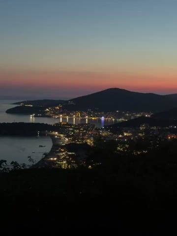 This is a vertical, medium-shot video taken from a high vantage point overlooking a coastal town at dusk. The sky displays a gradient of twilight colors, from a deep blue at the top to soft pink and orange hues near the horizon. Below the sky, a dark mountain range frames the scene, with the lights of the town twinkling across its slopes and along the coastline. The town itself appears to be situated around a bay, with a crescent-shaped beach visible in the foreground. The water of the bay is dark and calm, reflecting some of the distant lights. In the foreground, the dark silhouettes of trees and foliage obscure the very bottom of the frame, suggesting the viewer is looking out from a wooded area. The town lights create a warm, inviting glow against the darkening landscape. There are no people or specific objects clearly identifiable, just the expansive view of the town and its natural surroundings transitioning into night. The overall impression is one of serene beauty and the gentle onset of nighttime.