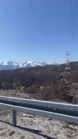 The video, shot from a moving vehicle, presents a clear sky over a mountainous landscape. Snow-capped mountain peaks are visible in the distance. The foreground includes sparse vegetation covering rolling hills. A red and white transmission tower stands on one of the hills. A silver guardrail runs along the edge of the road, positioned at the bottom of the frame. The road surface appears cracked and uneven. The overall scene is one of a rural, possibly mountainous, area on a sunny day.