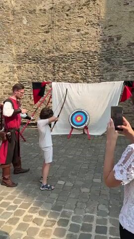 A boy in a light-colored t-shirt and shorts is aiming a bow and arrow at a target. He is standing on a cobblestone ground. To his left, a man dressed in a red tunic and brown boots, possibly an instructor, is observing him. To the right, a person's hand is visible holding a smartphone, possibly recording the event. The target is circular with concentric rings of blue, white, yellow, and red. A white cloth backdrop is behind the target, and a stone wall is visible further back. Red and black banners are attached to poles on the left side of the scene. The boy appears to be in the process of shooting or has just shot the arrow. The overall scene suggests an outdoor archery activity, possibly at a historical reenactment or festival.