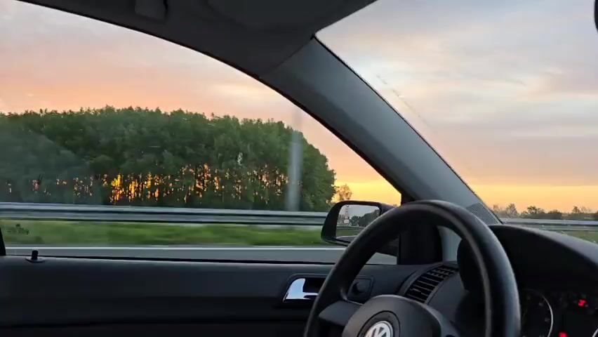 The video, taken from inside a moving car, shows a sunset over a highway. The sun is visible on the left side of the frame, partially obscured by trees. The car is moving at a high speed, and other vehicles can be seen passing by on the highway. The sky is a mix of orange, yellow, and blue, with some clouds. The windshield and car door are visible in the foreground, and the dashboard and steering wheel are partially visible on the right.