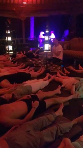 The video shows a room full of people lying on mats, seemingly relaxed. A man is sitting in the corner of the room near what appears to be some kind of singing bowl. The lighting in the room is dim and colored. The scene appears to be indoors, maybe in a yoga studio or similar space, with a wooden structure visible. The people are dressed in casual clothing.