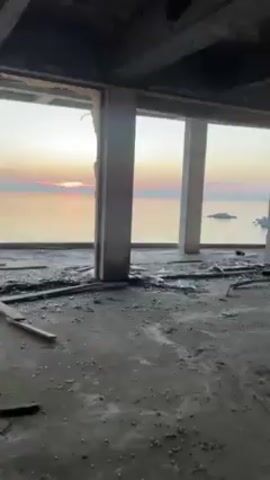 The camera pans around an unfinished or abandoned room, revealing a picturesque sunset over the ocean visible through large window openings. The room is mostly concrete with exposed beams and pillars. Debris litters the floor. The view from the window shows the ocean and a rocky islet. A rocky cliff face is also visible in the background.