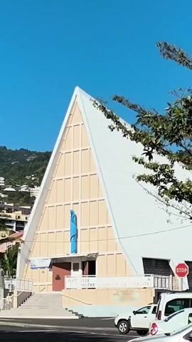 A still shot of a church with a distinctive triangular roof and a blue statue of Mary on its facade. A white SUV is parked in front of the church, and a few other cars are visible on the street. The scene is set against a clear blue sky and a lush green hillside dotted with houses. The overall impression is one of a peaceful, sunny day in a tropical or subtropical environment.