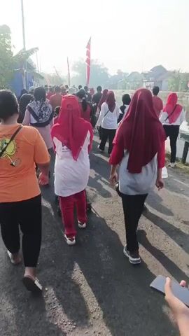 The video shows a group of people walking along a paved road under a bright, possibly morning, sky. The people are walking away from the camera. Most of them are wearing casual clothing, including t-shirts, leggings, and headscarves. Some individuals are wearing red and white outfits, possibly indicating a uniform or participation in an event. Several red and white flags are visible along the side of the road, suggesting a celebration or national holiday. The environment appears to be a semi-rural or suburban area, with some houses and vegetation visible in the background. The people are walking in the same direction, suggesting they are part of a procession or organized activity.