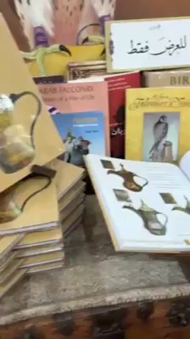 The video shows a display of books on falconry and desert culture, with an open book displaying illustrations of antique coffee pots. Behind the books, there is a table with various cultural items, including rugs, wooden bowls, and other decorative objects. A man in a black suit walks past the display. The environment appears to be a cultural exhibition or market.