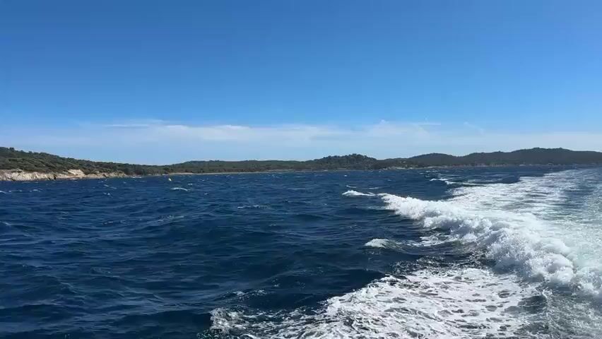 This media file shows a view from a boat moving through choppy, dark blue water. In the distance, a lush green coastline with rolling hills is visible under a bright, clear blue sky. The sun is high, indicating it's likely midday.

On the left side of the frame, the coastline is rocky and covered in dense, dark green trees. As the boat moves, the view shifts to reveal more of the coastline, which appears to be an island or a peninsula. A prominent feature is a stone structure, possibly a fort or an old building, situated on a hill overlooking the sea. The building is light brown and appears to be made of large stone blocks, with some sections looking weathered.

The sea is active, with white-capped waves and a visible wake trailing behind the boat, suggesting movement. The overall atmosphere is one of a bright, sunny day at sea, with the natural beauty of the coastline as the backdrop. There are no people or specific objects clearly identifiable in the water apart from the natural waves and the wake. The scene conveys a sense of travel and exploration of a scenic coastal area. The location is identified as Hyères, France, which is consistent with the Mediterranean coastal scenery depicted. No visible text is present in the media file.