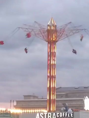 A tall, illuminated amusement park ride spins people in red seats against a cloudy sky. The ride is a vertical structure with arms extending outward, from which swings hang. The central tower is lit with vertical stripes of light. Below the ride, a building with lights and signage is visible. The sign reads "ASTRA COFFE." A person is visible near the signage. The overall atmosphere is that of an amusement park at dusk or on an overcast day.