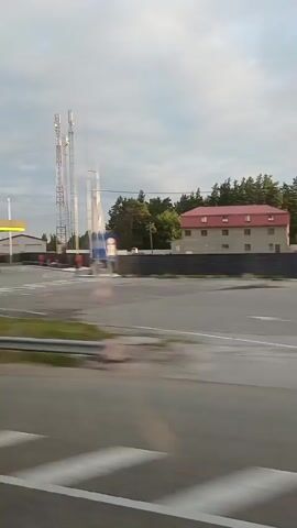 The video shows a moving vehicle's perspective, looking out a window. The scenery passes by a gas station with a yellow canopy and signage. There are cell towers in the background. A building with a red roof and white walls is visible, along with a large tree. Further down the road, more trees and a blue fence are seen. The sky is cloudy.