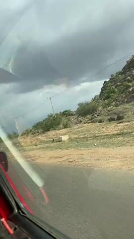 A car passenger films a view of an open field with a cloudy sky above. The car is red, and the video is taken from inside the vehicle. The landscape appears dry and contains some trees and rocks. Power lines are visible along the road. The sky is overcast with dark, heavy clouds, suggesting an imminent rainstorm. The road is paved, and the adjacent land is mostly dirt.