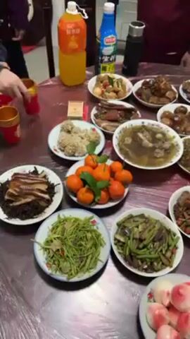 The video shows a large round table laden with a variety of Chinese dishes. There are multiple plates of food, including meat dishes, vegetables, soup, oranges, and peach-shaped buns. People are seated around the table, and one person uses a spoon to serve soup. There are also bottles of juice and a thermos on the table. The setting appears to be a home dining room.