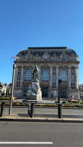 The video shows a statue in front of a large, ornate building. The statue appears to be of a man in historical clothing, standing on a pedestal with two female figures at its base. The building behind the statue is a grand structure with multiple floors, arched windows, and decorative architectural details. The sky above is clear and blue. The video is shot from a slightly elevated perspective, possibly from across a street or walkway. The camera is relatively still, but there is a slight wobble, suggesting it might be handheld. The scene is brightly lit by sunlight.