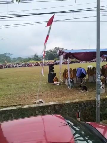 A medium-shot video pans across a grassy field where an outdoor event is taking place. In the foreground, utility poles and wires are visible against an overcast sky. A red car is partially in frame on the left. A dark-colored pickup truck is also in the foreground on the right.

In the mid-ground, people are gathered under a large maroon tent. Some individuals are seated in rows of chairs, while others stand. A woman in a light-colored dress and a headscarf is walking with a bouquet of white flowers. A tall flagpole with a red and white striped flag is visible. In the background, a crowd of people are spread across the field, and more tents are set up.

To the left of the tent, a portable speaker is positioned on the grass. A sign with Indonesian text is visible near the front of the field. The overall scene suggests a community gathering or ceremony. The weather appears to be overcast or possibly raining, as some people are holding umbrellas.