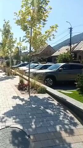 The video shows a sunny day with a clear blue sky. A modern building with dark brick and large glass windows is on the left side of the frame. In front of the building is a paved sidewalk made of light-colored rectangular stones. Beyond the sidewalk, there is a row of young trees planted in the ground. Several parked cars are visible behind a wooden fence bordering the trees. Further back, houses with pitched roofs can be seen. Three people are walking away from the camera on the sidewalk, in front of the modern building. The camera is panning slowly to the right.