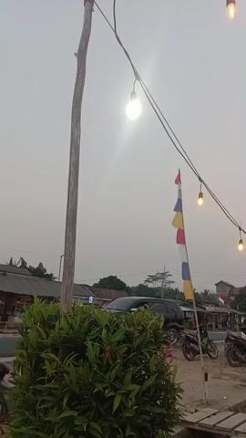 The video shows a string of lights hanging from a wooden pole. In the foreground, there is a dense green bush. In the background, there is a road with several motorcycles parked along it. There are also buildings and trees visible. The sky is overcast.