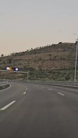 The video shows a view from a car driving on a highway. The highway has multiple lanes and guardrails on the sides. On the right side of the highway, there is a hill covered with dry grass and scattered trees. Above the highway, there is a large electronic sign with an image of a tunnel and text that reads "TUNEL DE MORATA 2". There are a few cars visible on the highway. The sky is a pale, hazy yellow, suggesting it might be early morning or late evening.