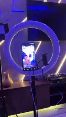 The video shows a living room with colorful lighting.  In the first shot, a smartphone is mounted in a ring light, showing a live video of a man.  The next shot pans around the room, showing a couch, a large television mounted on the wall, and various decorative lighting fixtures. The lighting changes from orange and green to blue.