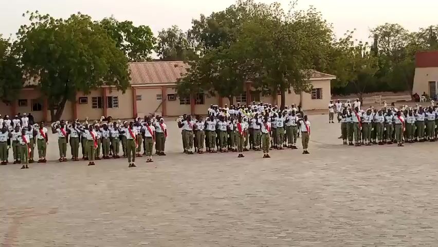 In Ajiwa, Nigeria, a large group of people, appearing to be young adults in uniform, are assembled in an outdoor area. The individuals are dressed in green pants and white shirts with red sashes draped over their shoulders. They stand in formation on a dusty, flat ground, with buildings and trees visible in the background. The buildings have a light-colored facade with numerous windows, suggesting a school or training facility. Trees with dense green foliage are scattered around the perimeter.

The people are engaged in a drill or ceremony, as indicated by their organized rows and synchronized movements. At one point, many of them raise their right hands to their foreheads in a salute, a common gesture in military or cadet activities. The overall atmosphere appears formal and disciplined, with no overt expressions of emotion visible.

The time of day seems to be late afternoon or early evening, given the soft, diffused light and the absence of harsh shadows. The sky is a pale, hazy color, hinting at a warm and possibly dry climate. The presence of a large, open space and the uniform attire suggest this is an organized event, possibly a training exercise, parade, or ceremonial gathering. There is no visible text in the media file.