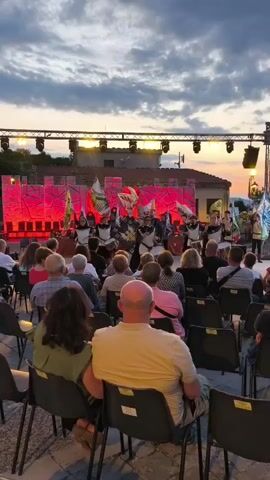 A vertical video shows an outdoor performance at dusk. In the foreground, a crowd of people sits in rows of black chairs, facing a stage. The stage is set with a red backdrop and illuminated by red lights. On stage, several performers dressed as medieval knights stand holding flags. The sky above is cloudy with hints of orange and pink from the setting sun. There is a sound of drums and fanfare.
