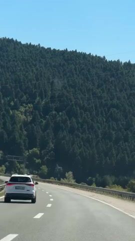 The video shows a view from a vehicle driving on a highway. The highway curves to the right. On the left side of the highway, there is a dense forest covering a hillside. Several cars are visible on the highway, including a white car in the foreground and a white car further ahead. There are also overhead signs and barriers along the side of the road. The sky is clear and blue.