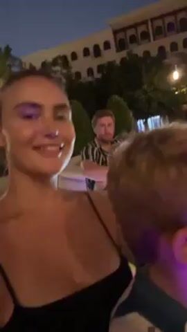 A woman films herself and two young boys in the back of a ride-on cart. The cart, operated by a man in the front seat, is lit up with a variety of glowing lights. The video is shot at night; in the background is a body of water with fountains. The woman smiles and laughs.