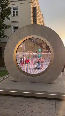 A person's hand with a ring and a watch is visible in the foreground, reaching towards the right side of the frame. In the middle ground, a large circular sculpture with a digital display is positioned. The display shows a scene with a red scooter and several people walking on a street, with buildings and traffic lights in the background. The sun appears to be setting, casting a warm glow on the scene. The ground is paved with tiles, and a strip of lights is visible at the base of the sculpture.