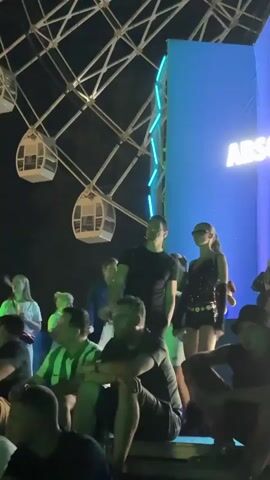 The video shows a group of people gathered outdoors at night. A Ferris wheel with illuminated cabins is visible in the background. To the right, a large blue inflatable structure displays the Absolut logo. In the foreground, several individuals are seated or standing, some appearing to be watching a performance or event. The lighting shifts from ambient night illumination to red lights, suggesting a dynamic atmosphere. One woman in the center appears to be a focal point, dressed in a sparkly outfit and sunglasses.