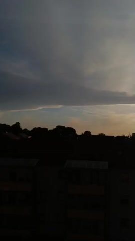 A vertical video shows a city skyline at dusk. The sky is filled with dramatic clouds, ranging from dark gray to a pale orange as the sun sets. Several contrails are visible, forming interesting patterns in the sky. Silhouetted against the sky are the rooftops of buildings, trees, and the outlines of some taller structures in the distance. In the foreground, the dark silhouette of an apartment building takes up the lower portion of the frame. The lighting is dim, characteristic of twilight, with the setting sun casting a warm glow on the horizon.