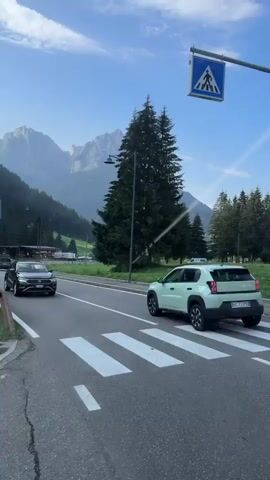 The video shows a mountain road with two cars, a dark colored SUV and a light green SUV, driving past a crosswalk. In the background, there are trees, mountains, and a blue sky with some clouds. The road is paved and has white lines marking the lanes and the crosswalk. There are also pedestrian crossing signs visible. The overall scene is serene and depicts a clear day in a mountainous region.